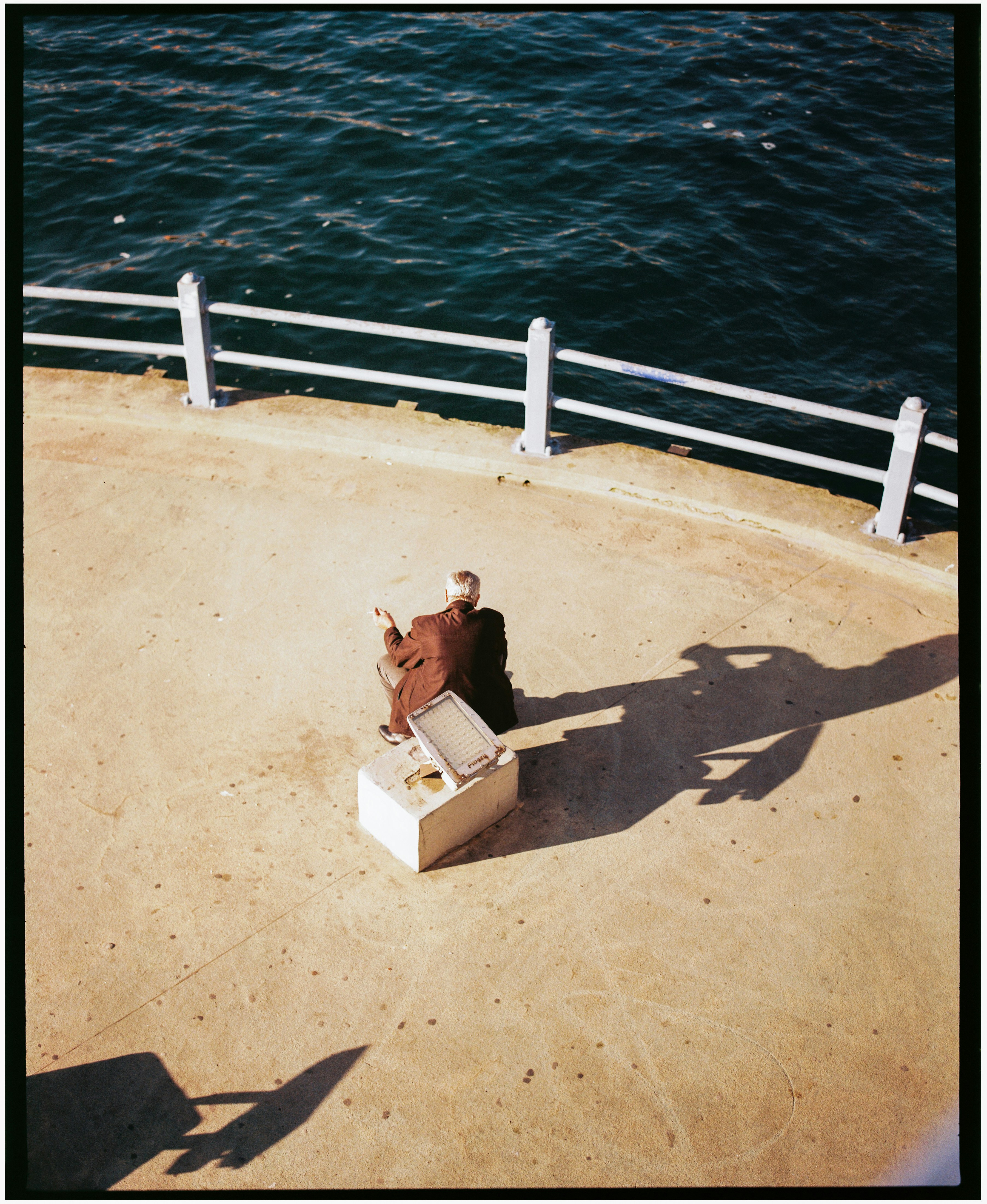 An adult in a brown jacket sitting on a concrete block by a waterfront railing