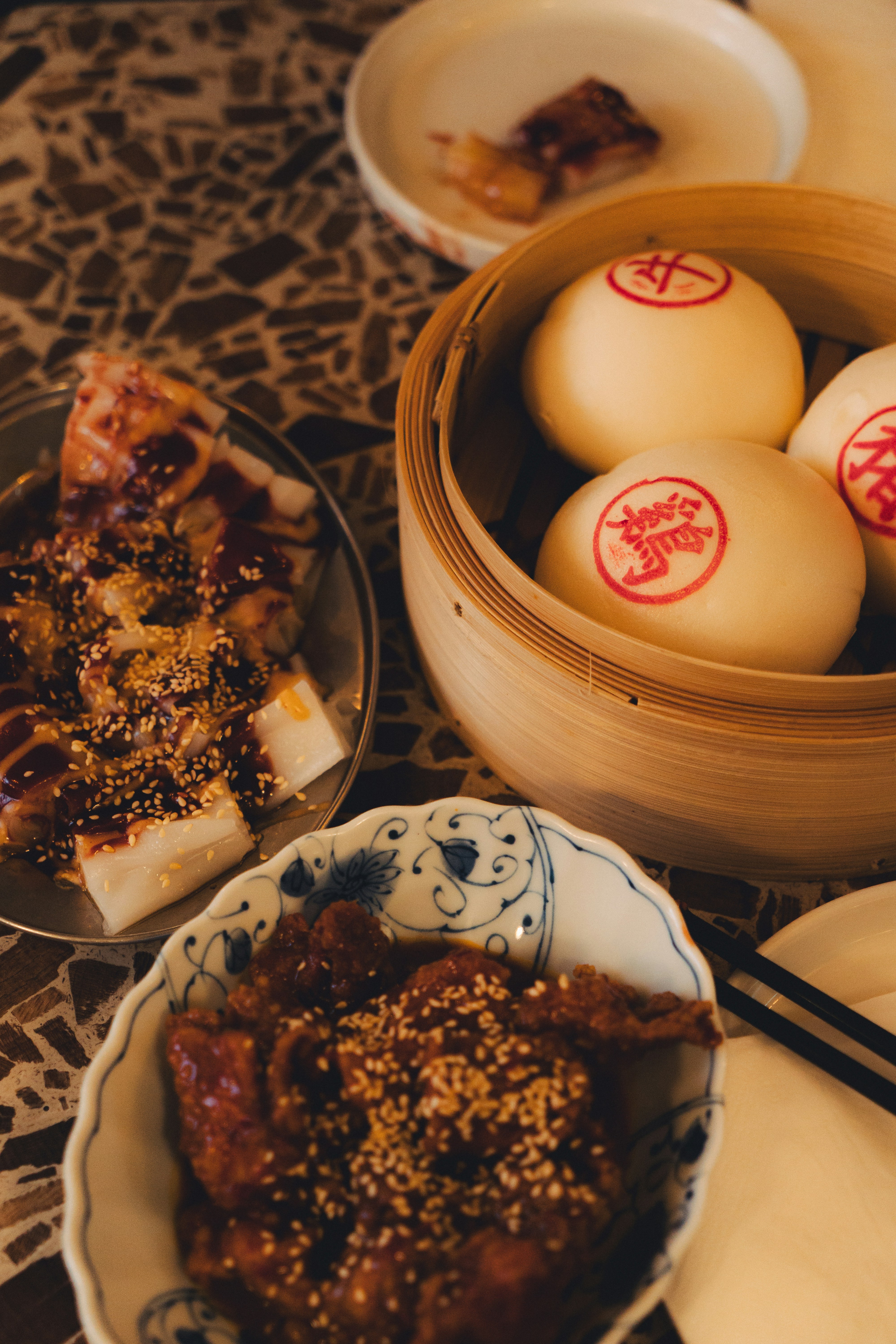 Steamed buns in a bamboo basket beside plates of dim sum on a patterned table