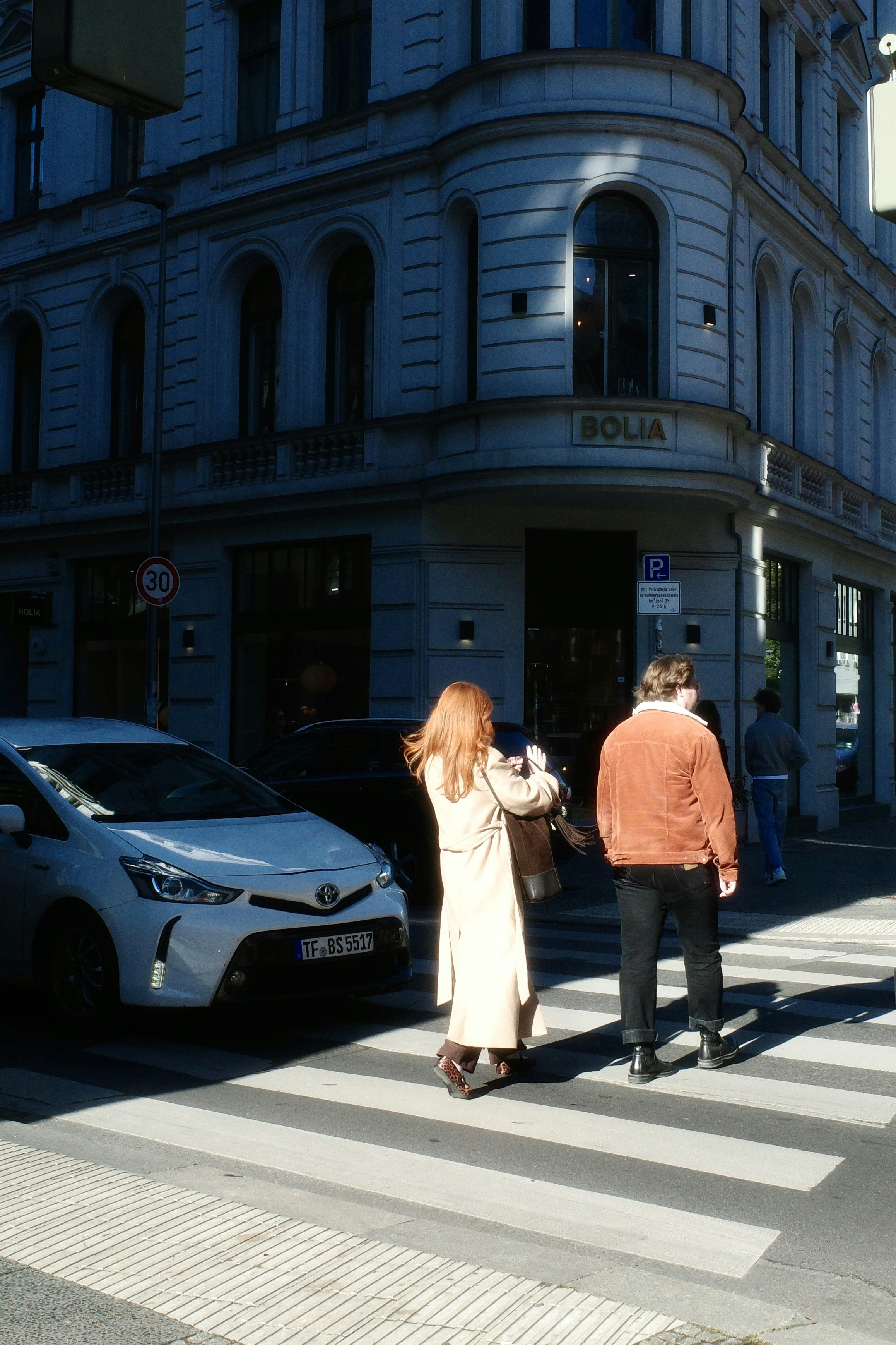 Two people crossing a city street in front of a white car near a corner building - 相关推荐
