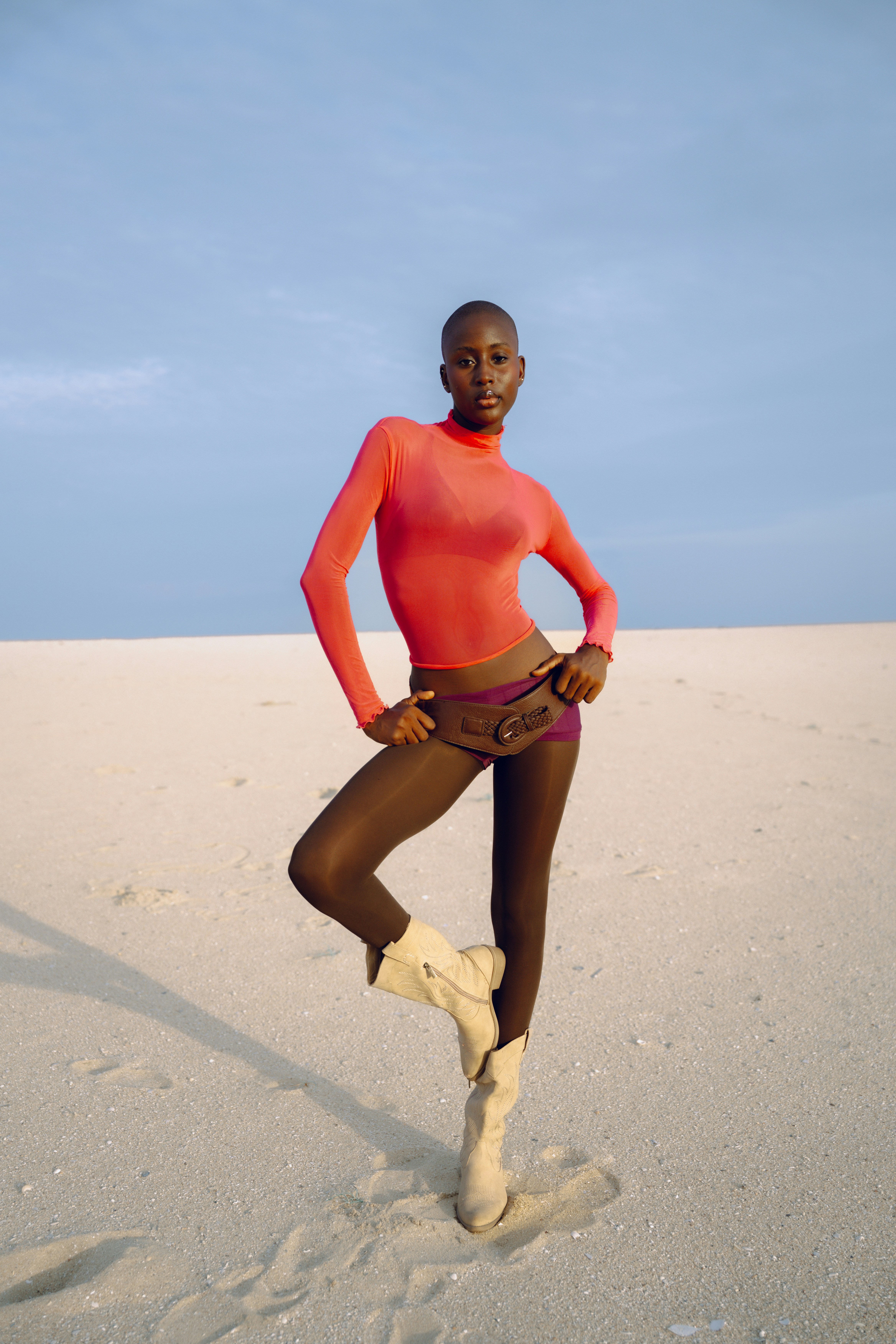 A person with a shaved head in a bright orange top and brown leggings on a sandy beach