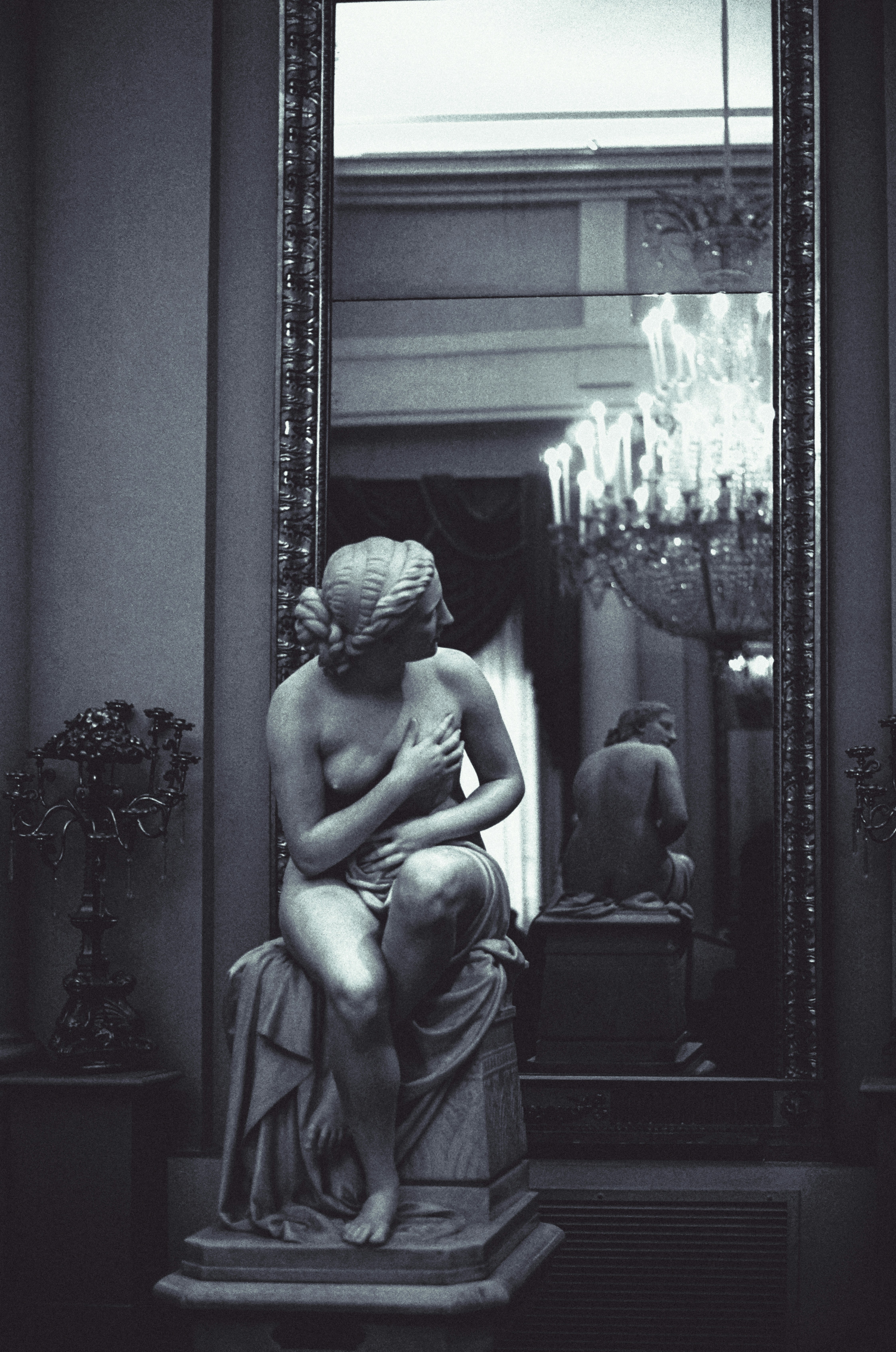 A marble statue of a seated woman reflected in a large ornate mirror with a chandelier