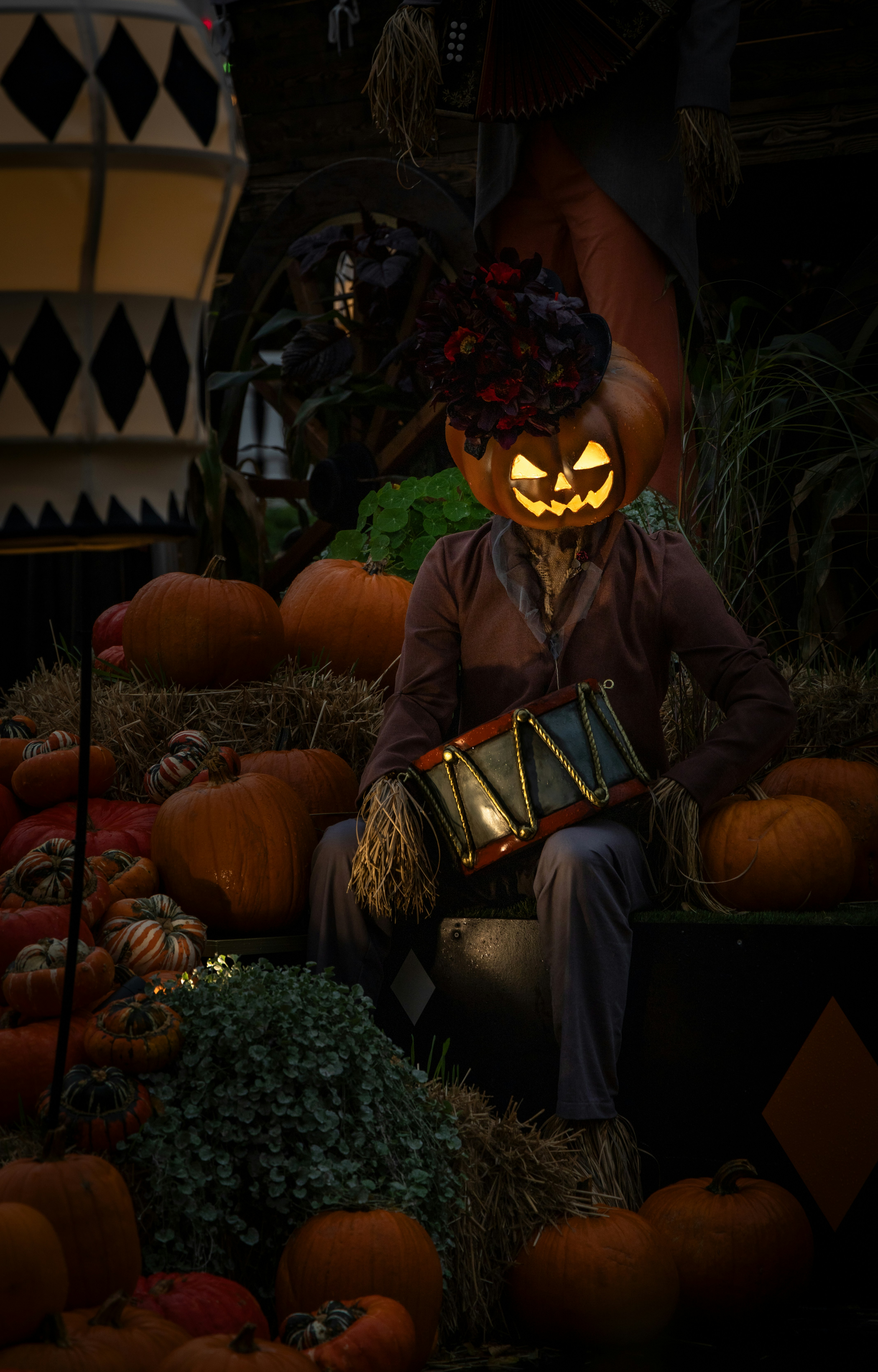 A scarecrow with a glowing jack-o'-lantern head sitting among many pumpkins on hay bales