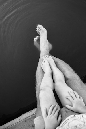 A black and white photograph of a person with a child sitting on their lap. Both individuals are dipping their feet into calm water, creating gentle ripples. The focus is on the legs and feet, emphasizing the relaxed atmosphere.