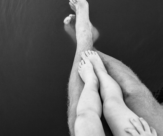A black and white photograph of a person with a child sitting on their lap. Both individuals are dipping their feet into calm water, creating gentle ripples. The focus is on the legs and feet, emphasizing the relaxed atmosphere.