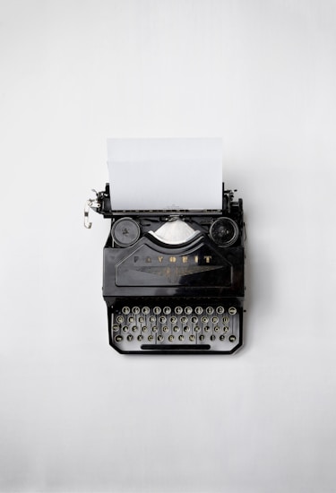 An old-fashioned black typewriter sits on a light grey background. The typewriter has a piece of blank paper inserted into it. The keys of the typewriter are visible along with some mechanical components.