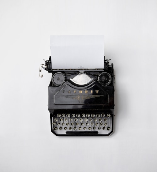 An old-fashioned black typewriter sits on a light grey background. The typewriter has a piece of blank paper inserted into it. The keys of the typewriter are visible along with some mechanical components.