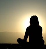 A serene meditation scene with a woman sitting cross-legged by a calm lake at sunrise.
