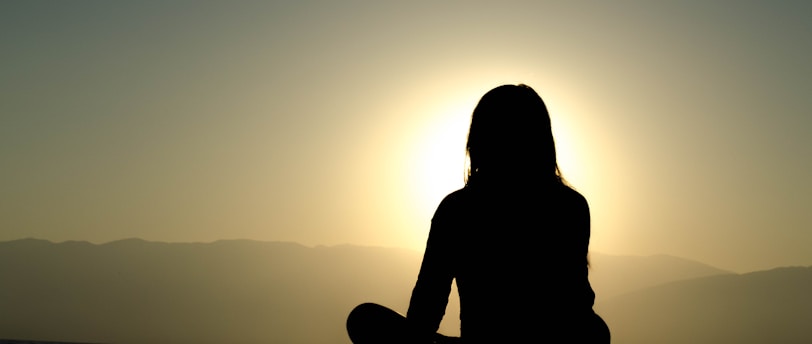 A silhouette of a person sitting cross-legged with the backdrop of a setting or rising sun, creating a halo effect around the head and shoulders. The scene is serene with calm water and distant mountains partially visible in the background.