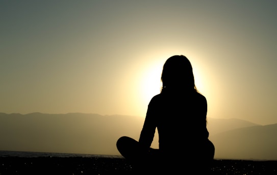A silhouette of a person sitting cross-legged with the backdrop of a setting or rising sun, creating a halo effect around the head and shoulders. The scene is serene with calm water and distant mountains partially visible in the background.