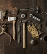 A collection of vintage woodworking tools arranged neatly on a rustic wooden surface. The tools include various hammers, an axe, gloves, a hand saw, scissors, a metal cup filled with dark liquid, and other hand tools. The overall composition suggests an antique or workshop setting.