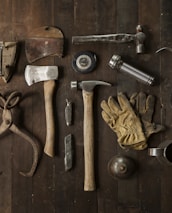 A collection of vintage woodworking tools arranged neatly on a rustic wooden surface. The tools include various hammers, an axe, gloves, a hand saw, scissors, a metal cup filled with dark liquid, and other hand tools. The overall composition suggests an antique or workshop setting.
