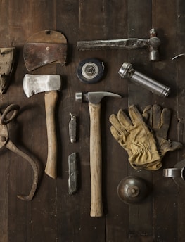 A collection of vintage woodworking tools arranged neatly on a rustic wooden surface. The tools include various hammers, an axe, gloves, a hand saw, scissors, a metal cup filled with dark liquid, and other hand tools. The overall composition suggests an antique or workshop setting.
