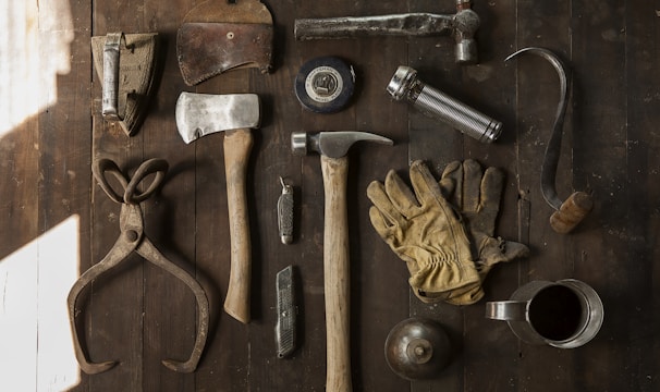 A collection of vintage woodworking tools arranged neatly on a rustic wooden surface. The tools include various hammers, an axe, gloves, a hand saw, scissors, a metal cup filled with dark liquid, and other hand tools. The overall composition suggests an antique or workshop setting.