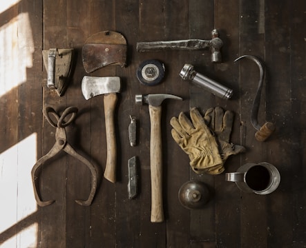 A collection of vintage woodworking tools arranged neatly on a rustic wooden surface. The tools include various hammers, an axe, gloves, a hand saw, scissors, a metal cup filled with dark liquid, and other hand tools. The overall composition suggests an antique or workshop setting.