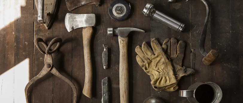 Vintage tools including an axe, hammer, gloves, and metal implements on dark wooden planks