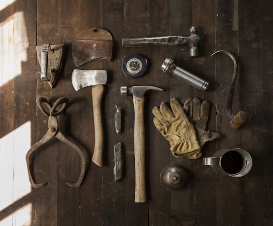 A collection of vintage woodworking tools arranged neatly on a rustic wooden surface. The tools include various hammers, an axe, gloves, a hand saw, scissors, a metal cup filled with dark liquid, and other hand tools. The overall composition suggests an antique or workshop setting.