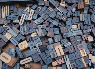 A collection of vintage wooden letterpress blocks arranged in a seemingly random pattern. The blocks feature a variety of fonts, sizes, and orientations, showcasing letters and numbers in a mix of light and dark shades.