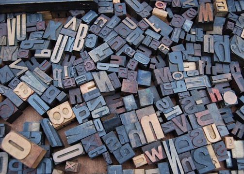 A collection of vintage wooden letterpress blocks arranged in a seemingly random pattern. The blocks feature a variety of fonts, sizes, and orientations, showcasing letters and numbers in a mix of light and dark shades.