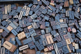 A collection of vintage wooden letterpress blocks arranged in a seemingly random pattern. The blocks feature a variety of fonts, sizes, and orientations, showcasing letters and numbers in a mix of light and dark shades.