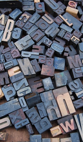 A collection of vintage wooden letterpress blocks arranged in a seemingly random pattern. The blocks feature a variety of fonts, sizes, and orientations, showcasing letters and numbers in a mix of light and dark shades.