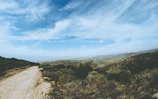 Wide dirt road stretching through rolling hills with scattered trees and distant mountains.