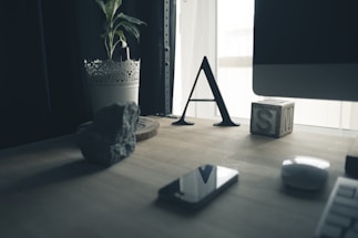 A wooden desk is adorned with various items including a potted plant in a laced metal holder, a decorative rock, a black letter 'A', a wooden block with the letter 'S' on it, a smartphone, and a computer mouse. A portion of a computer screen is partially visible on the right side. The window in the background lets in natural light, creating a soft atmosphere.