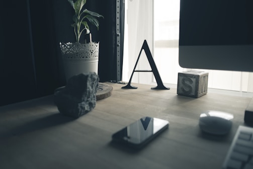 A wooden desk is adorned with various items including a potted plant in a laced metal holder, a decorative rock, a black letter 'A', a wooden block with the letter 'S' on it, a smartphone, and a computer mouse. A portion of a computer screen is partially visible on the right side. The window in the background lets in natural light, creating a soft atmosphere.