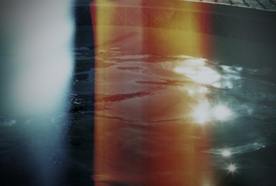 A photograph featuring an artistic play of light leaks over a surface of water, presumably a pool. The light leaks vary in colors, notably yellow, orange, and blue. The water surface shows reflection and light flares, adding a dreamy and surreal effect to the image. The tiles around the edge of the pool are partially visible, indicating location context.