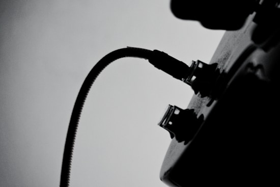 A black and white silhouette of a cable plugged into a device with visible knobs and a socket. The image focuses on the curved cable, creating a contrast against the light background.