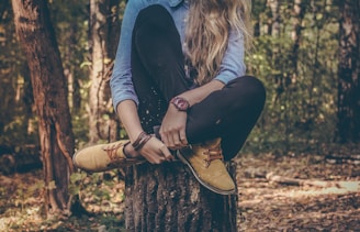 A person with long hair sits cross-legged on a tree stump in a forest. They are wearing a denim shirt, black pants, and yellow boots, and have a red watch on their left wrist. The background is filled with trees and foliage.