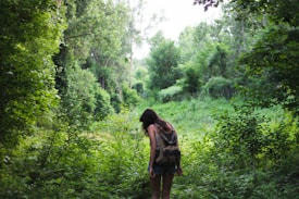 A person with long dark hair is walking through a lush, green forest. They are carrying a brown backpack and appear to be immersed in the natural surroundings. Tall trees and dense greenery create a serene and peaceful environment.