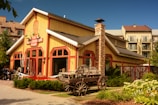A rustic wooden store building with a red and yellow exterior featuring a large stone chimney sits amidst a green landscape. In front of the building, there is a wooden cart and some outdoor seating is visible where a few people are sitting. The background includes a larger residential or hotel building with balconies.