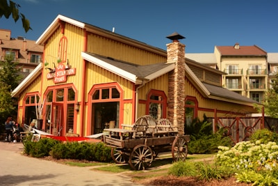 A rustic wooden store building with a red and yellow exterior featuring a large stone chimney sits amidst a green landscape. In front of the building, there is a wooden cart and some outdoor seating is visible where a few people are sitting. The background includes a larger residential or hotel building with balconies.