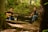 Two men are seated in a wooded area, each playing an acoustic guitar. They appear relaxed and engaged in music, surrounded by trees and lush greenery. A guitar case is visible on the ground in front of them.