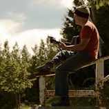 A person wearing a beanie and an orange shirt is playing a guitar while sitting on a rusty metal structure. The background features lush greenery and clear skies, suggesting a serene outdoor setting.