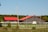 Two buildings with striking red roofs are situated within a fenced grassy area, set against a backdrop of dense green trees. Wooden posts and a white fence enclose the area, and there's a clear blue sky overhead.
