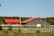 Two buildings with striking red roofs are situated within a fenced grassy area, set against a backdrop of dense green trees. Wooden posts and a white fence enclose the area, and there's a clear blue sky overhead.