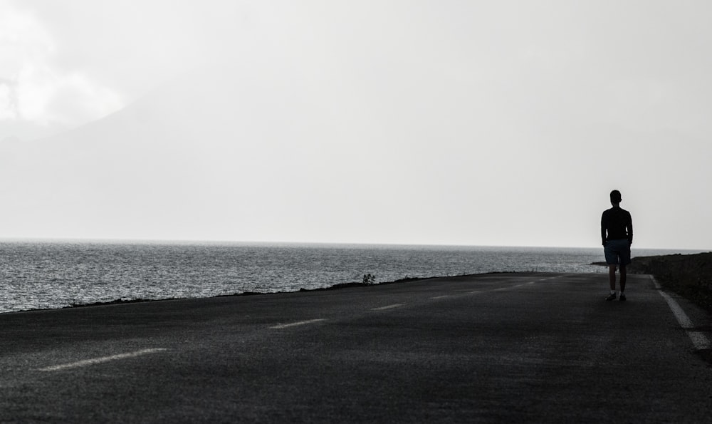 man standing on asphalt road beside body of water under gray sky