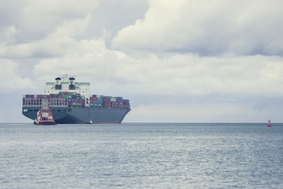 A large cargo ship loaded with numerous colorful shipping containers is floating on the calm sea. A smaller tugboat is positioned near the forefront of the large vessel, assisting with navigation. The sky is filled with thick, gray clouds, suggesting the possibility of overcast weather.