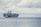 A large cargo ship loaded with numerous colorful shipping containers is floating on the calm sea. A smaller tugboat is positioned near the forefront of the large vessel, assisting with navigation. The sky is filled with thick, gray clouds, suggesting the possibility of overcast weather.