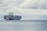A large cargo ship loaded with numerous colorful shipping containers is floating on the calm sea. A smaller tugboat is positioned near the forefront of the large vessel, assisting with navigation. The sky is filled with thick, gray clouds, suggesting the possibility of overcast weather.