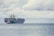 A large cargo ship loaded with numerous colorful shipping containers is floating on the calm sea. A smaller tugboat is positioned near the forefront of the large vessel, assisting with navigation. The sky is filled with thick, gray clouds, suggesting the possibility of overcast weather.