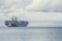 A large cargo ship loaded with numerous colorful shipping containers is floating on the calm sea. A smaller tugboat is positioned near the forefront of the large vessel, assisting with navigation. The sky is filled with thick, gray clouds, suggesting the possibility of overcast weather.