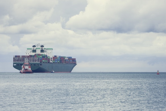 A large cargo ship loaded with numerous colorful shipping containers is floating on the calm sea. A smaller tugboat is positioned near the forefront of the large vessel, assisting with navigation. The sky is filled with thick, gray clouds, suggesting the possibility of overcast weather.