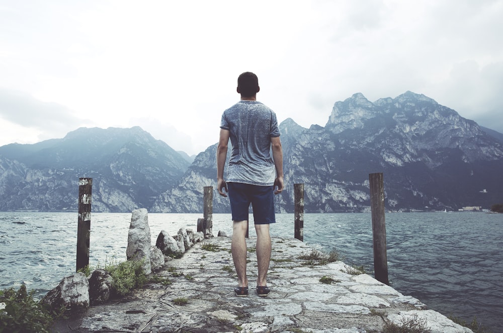 uomo in piedi sul molo di cemento grigio di fronte allo specchio d'acqua e alle montagne durante il giorno