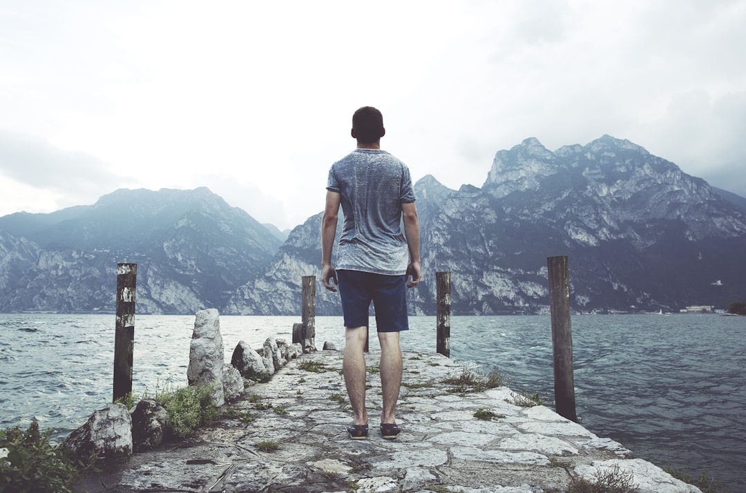 man standing on gray concrete dock facing body of water and mountains at daytime, Stone jetty on a cloudy day