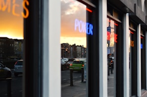 Reflections of a street with moving cars seen on the windows of a building at sunset. Neon lights with words like 'POKER' and other indistinguishable text are reflected on the glass. Buildings and traffic lights are visible in the background under a golden-orange sky.