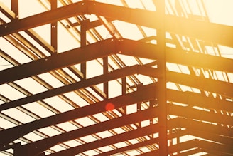 View of a construction framework with steel beams forming geometric patterns, captured at an angle with warm sunlight streaming through, creating a lens flare.