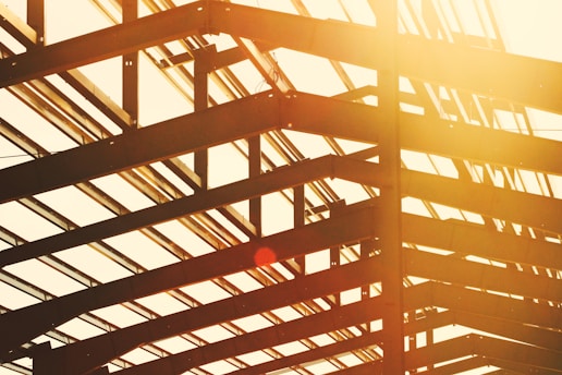 View of a construction framework with steel beams forming geometric patterns, captured at an angle with warm sunlight streaming through, creating a lens flare.