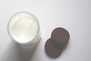 A glass of milk is placed on a smooth, light-colored surface next to two chocolate cookies. The milk is in a translucent glass, and the cookies have detailed circular patterns on them.